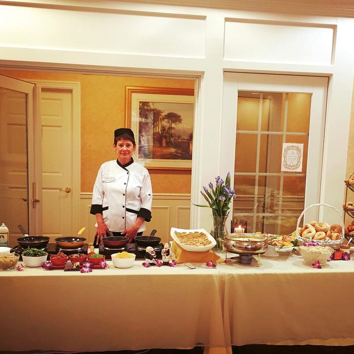 Chef Antoinette in her chef's coat behind a live catering station of sauté pans, garnishes and floral arrangements at an event